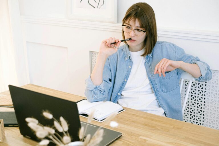 Woman sitting on a desk, with her pen on her mouth while looking at her notebook