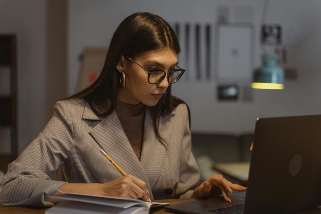 Woman writing down notes while scrolling on her laptop