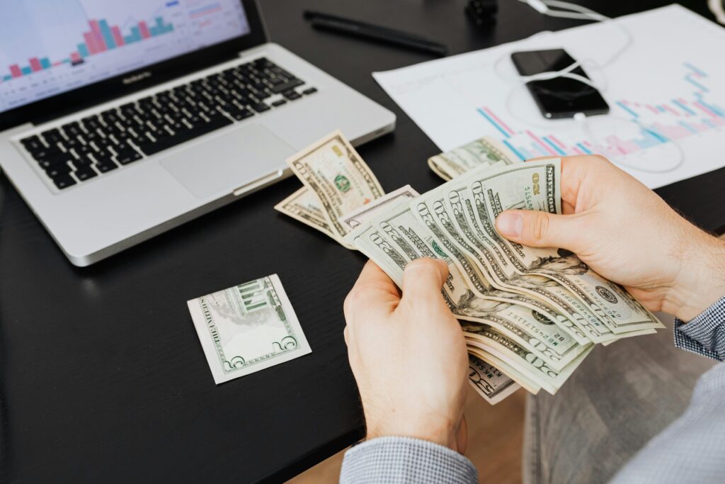 Person counting dollar bills while seated in front of his laptop